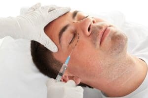 A male with short hair is undergoing a hyaluronic acid injection below his left eye by a plastic surgeon.